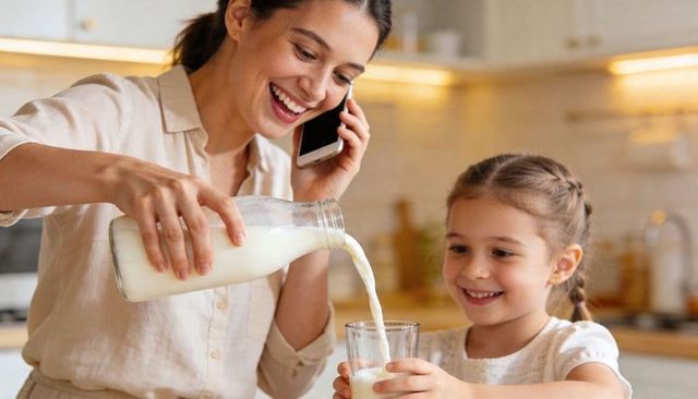 Smiling mother pouring milk for daughter while talking on phone in cozy kitchen morning