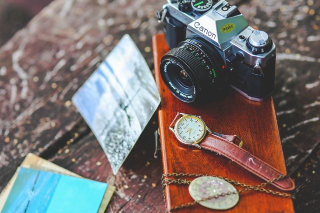 A vintage film camera, leather watch, and old photographs on a wooden table. Perfect for themes related to nostalgia, history, photography, and traveling back in time. Ideal for use in articles or advertisements about classic photography equipment, nostalgic moments, or vintage accessories.