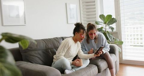Two friends sharing self-care morning with under-eye patches on cozy sofa, browsing phone