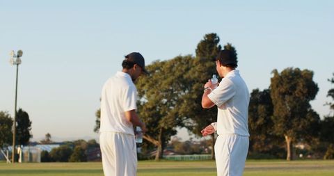 Diverse cricketers drinking water on grass during match break wearing white kit