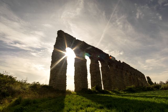 Photo depicting ancient Roman aqueduct ruins at sunset, with beams of sunlight streaming through the archways. Ideal for use in historical content, travel blogs, educational materials on architecture, or as a scenic backdrop for design projects focusing on history or ancient civilizations.