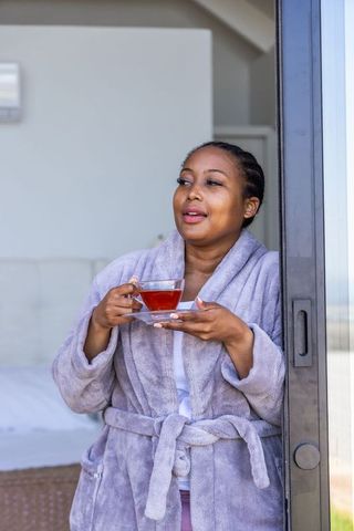 African american woman enjoying morning tea in lavender robe at sunlit bedroom doorway