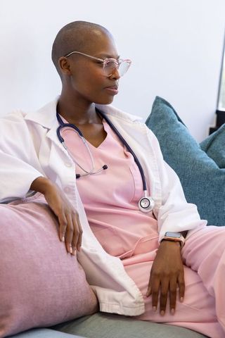 African female clinician relaxing on sofa wearing pink scrubs, white coat, stethoscope