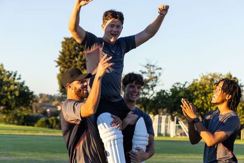Teammates lifting cricketer, celebrating victory and cheering on sunlit cricket field