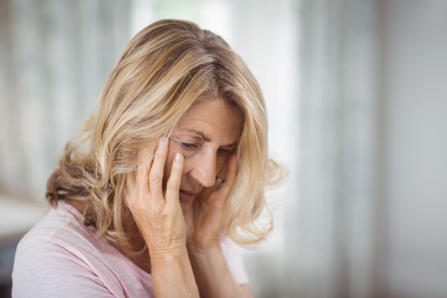 Senior woman sitting in bedroom, looking tense and worried. She is holding her head with both hands, appearing deep in thought. This image can be used to depict themes of mental health, anxiety, stress, solitude, and the challenges faced by elderly individuals. Suitable for articles, blogs, and campaigns focused on senior care, mental wellness, and lifestyle.