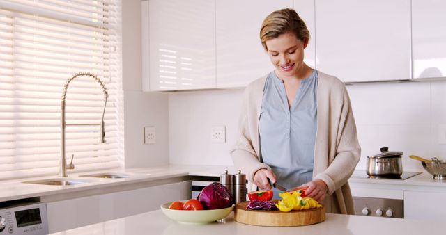 This scene is perfect for use in articles or advertising centered on healthy living, home cooking, kitchen design, or lifestyle choices. It depicts a woman in casual clothing smiling while she chops vegetables, set in a bright, modern kitchen. This can be used to promote kitchen appliances, cooking classes, healthy eating initiatives, lifestyle blogs, and magazines focused on home and family life.