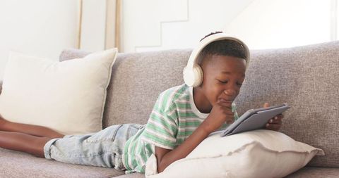 African boy lying on sofa wearing headphones using tablet for remote learning
