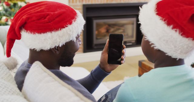 Father and son wearing Santa hats video calling using smartphone near fireplace, enjoying holiday time together. Perfect for holiday greeting cards, Christmas promotions, family technology use, festive bonding moments. Shows warmth, family connection, and use of modern technology during celebrations.