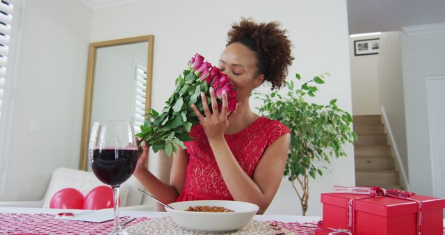 Image shows a woman in a red dress holding and smelling a bouquet of roses while sitting at a dinner table with a glass of red wine and neatly wrapped gift boxes. Ideal for use in advertisements, magazine covers, Valentine's Day marketing materials, or promotional banners for romantic events and gifts.