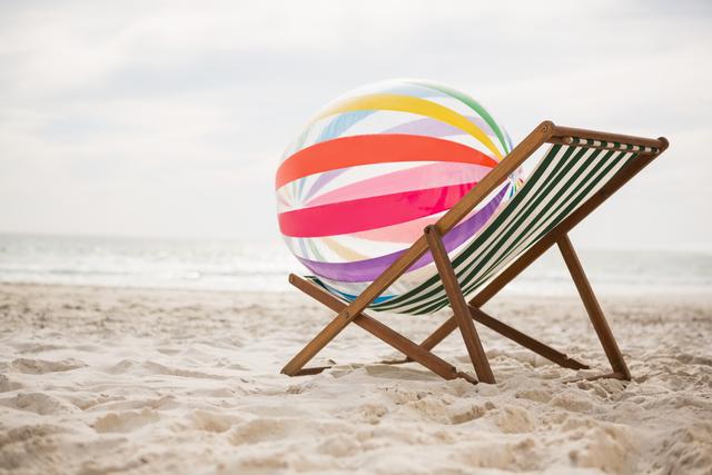 Colorful striped beach ball placed on an empty beach chair at a sandy tropical beach with the sea in the background. Ideal for vacation, travel, summer, and leisure concepts. Perfect for promoting beach holidays, travel agencies, relaxation products, and coastal lifestyle content.