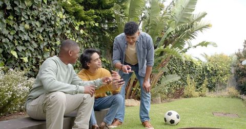 Three friends sharing tablet and laughing in lush backyard during casual soccer break