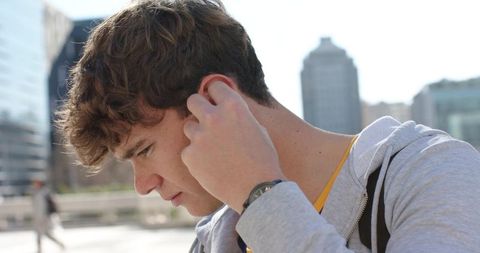 Young man adjusting earbud on urban plaza wearing hoodie, backpack and smartwatch daylight