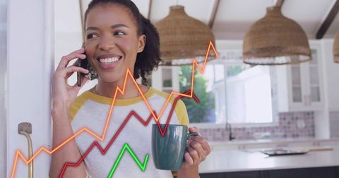Smiling woman talking on phone holding coffee mug in bright kitchen with rising graphs