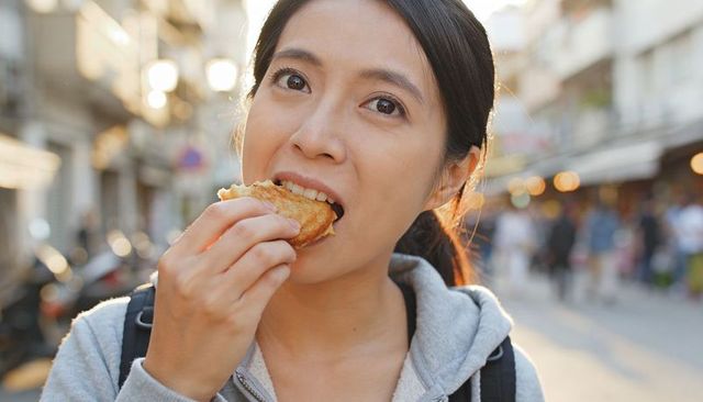 Young asian woman eating flaky pastry on bustling market street during golden hour