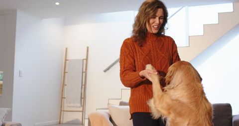 Woman wearing orange sweater holding golden retriever paws smiling in bright living room