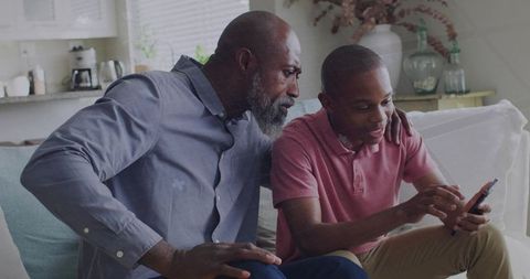 Father guiding son using smartphone on sofa in cozy daylight living room parenting moment