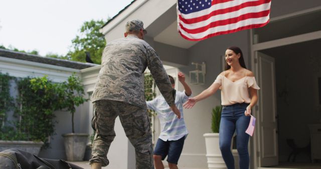 Happy caucasian male soldier lifting son and greeting wife with flag hanging outside their house. soldier returning home to family.