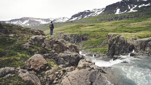 The image depicts a hiker exploring rocky mountain terrain with a flowing river. Green hills, rocky mountains, and patches of snow create a picturesque landscape. Ideal for use in travel blogs, adventure planning, and campaigns promoting outdoor activities and nature exploration.