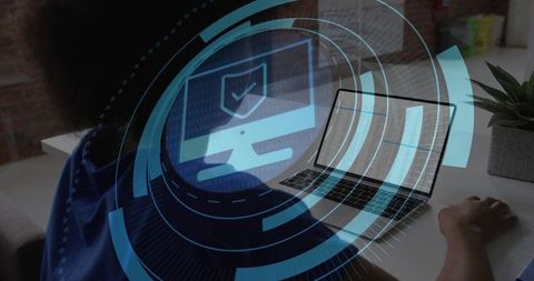 Curly-haired woman typing on laptop watching cybersecurity shield HUD overlay in modern workspace