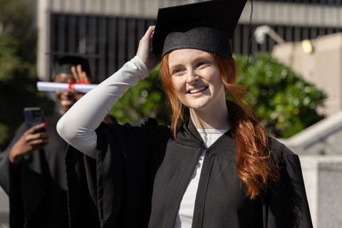 Joyful red-haired graduate adjusting mortarboard on campus steps holding diploma