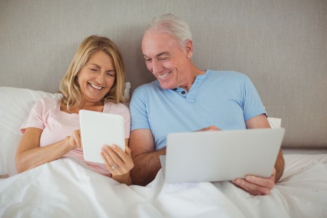 Senior couple enjoying time together in bed, using digital devices. Ideal for topics on senior lifestyle, technology use among elderly, and home leisure activities.