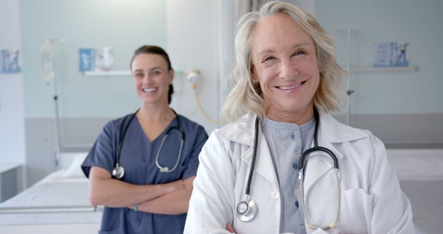 Two female medical professionals, a senior doctor and a nurse, are smiling warmly in a modern healthcare environment. The senior doctor, in a white lab coat with a stethoscope around her neck, stands in the foreground while the nurse in blue scrubs stands behind with arms folded. This image can be used in healthcare advertisements, medical websites, patient care brochures, or teamwork and collaboration campaigns in medical fields.