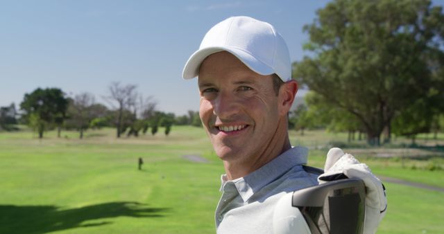 Adult male wearing a white cap holding a golf club over his shoulder, standing on a green golf course with trees in the background. He is smiling and looking at the camera, indicating enjoyment and relaxation. The scene denotes leisurely outdoor activity on a sunny day. Ideal for use in promotions related to golf, sports, outdoor recreation, hobbies, and leisure activities.