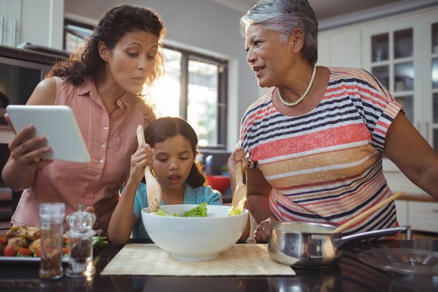 Three generations of women are preparing a meal together in a bright and modern kitchen. The grandmother, mother, and daughter are engaged in making a salad, with the mother holding a tablet, possibly following a recipe. This image is perfect for illustrating family bonding, healthy eating, and domestic life. It can be used in advertisements, blogs, and articles related to family activities, cooking, and lifestyle.