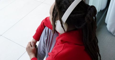 African american man listening with white headphones in red hoodie by bright window, relaxed