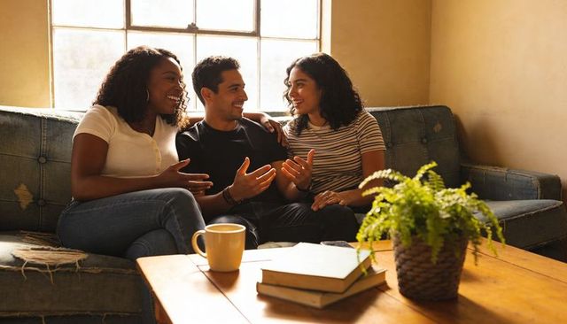 Friends chatting and laughing on worn blue sofa in cozy sunlit living room