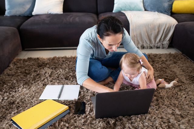 Caucasian mother multitasking by working on a laptop while caring for her baby daughter in a cozy living room. Ideal for articles or advertisements on remote work, parenting, work-life balance, and family life. Can be used in blogs, social media posts, or websites focusing on motherhood, home office setups, and modern family dynamics.