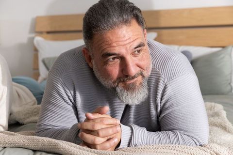 Mature man relaxing on bed in cozy neutral bedroom wearing light gray ribbed sweater