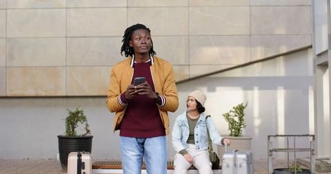 Young travelers waiting with luggage and using smartphone at modern urban plaza