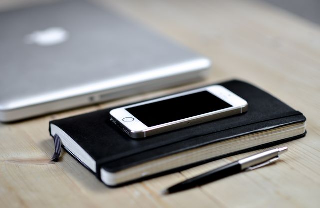 Smartphone lying on top of a leather notebook with a ballpoint pen beside it and a laptop in the background on a wooden desk. Ideal for topics related to technology usage, workplace productivity, study environment, office setup, and digital communication.