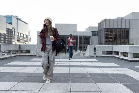 Young woman walking and talking on phone holding coffee on urban campus walkway