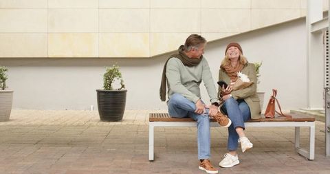 Mature couple laughing while sitting on bench in urban courtyard, casual autumn fashion