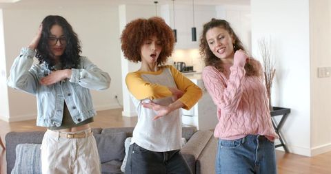 Three diverse women dancing and laughing in modern open-plan living room with kitchen