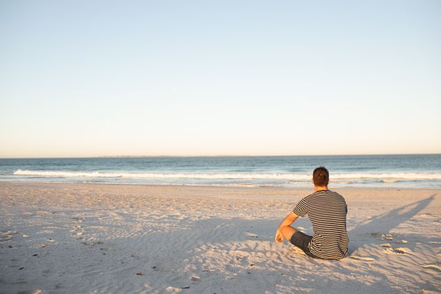 Man sitting on sandy beach during sunset, looking at ocean horizon. Ideal for themes of relaxation, solitude, nature, summer vacations, and peaceful moments. Suitable for travel blogs, wellness articles, and promotional materials for beach resorts.