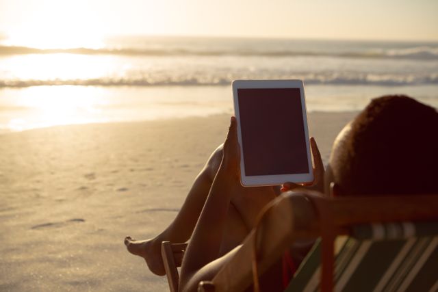 Woman relaxing on a beach chair while using a digital tablet during sunset. Ideal for themes related to technology use on vacation, leisure activities, summer lifestyles, and outdoor relaxation. Can be used in advertisements for beach resorts, travel agencies, digital devices, and lifestyle blogs.