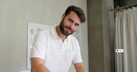 Bearded clinician leaning over exam table smiling in white polo clinical office