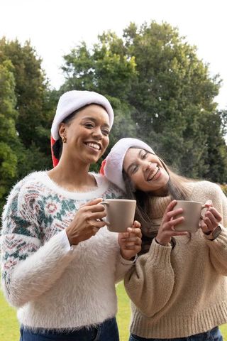 Diverse friends wearing santa hats holding steaming mugs in cozy winter park