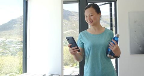 Asian woman smiling and checking smartphone while holding water bottle near window at home