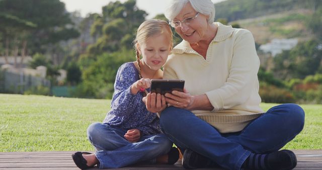 Elderly woman and young girl sitting on grass sharing fun moments using a tablet. Ideal for themes of family bonds, technology bridging generations, outdoor activities, and intergenerational learning.