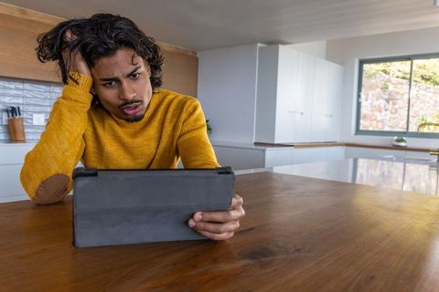 Young man wearing mustard sweater concentrating on tablet at modern kitchen table
