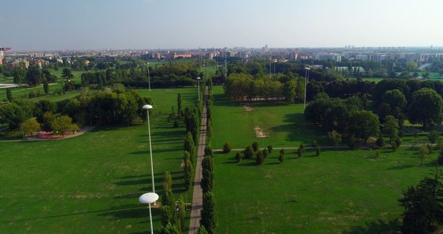 This photograph shows an expansive aerial view of a large urban park characterized by green fields, rows of trees, and an array of open spaces. The park is juxtaposed against a backdrop of a city skyline visible in the distance. This image can be used for travel brochures, urban development presentations, environmental projects, and websites promoting outdoor activities and recreation.
