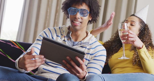 Couple sitting on couch, celebrating at home, watching tablet together. Man wearing glasses, woman drinking champagne with party hat. Perfect for themes on celebrations, modern lifestyle, digital engagement, home entertainment, and cozy environments.