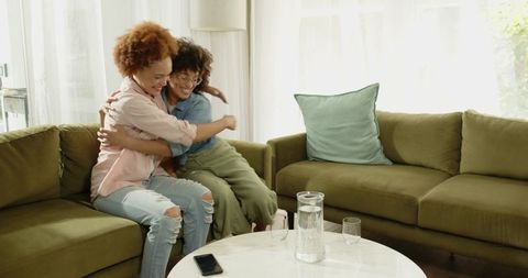 Two friends hugging and smiling on olive-green sectional sofa in sunlit living room