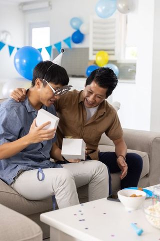 Asian father and son opening gift box on sofa during home birthday celebration