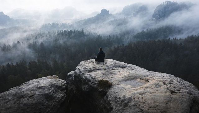 Solitary hiker sitting on rocky cliff overlooking misty pine valley and distant peaks