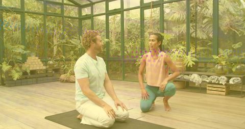 Yoga instructor teaching kneeling pose with student in greenhouse studio with plants